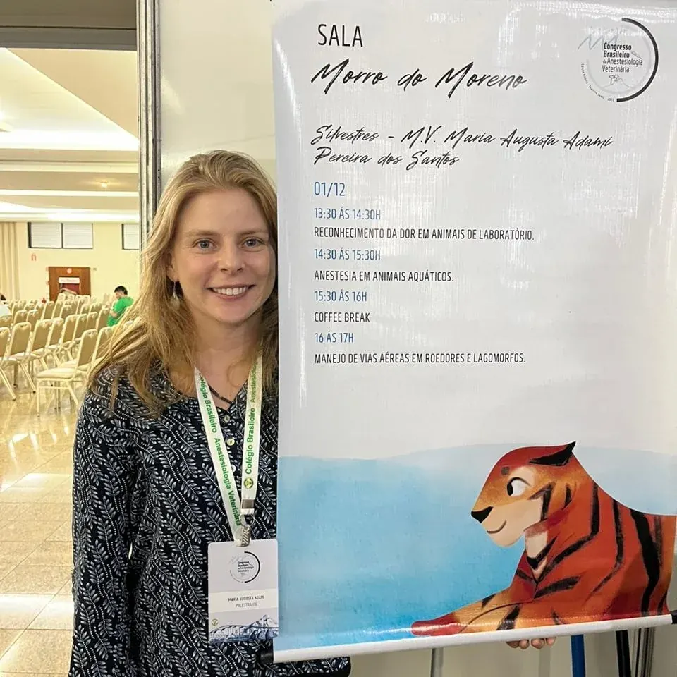 Guta com crachá de evento ao lado do painel da sala com a programação das palestras