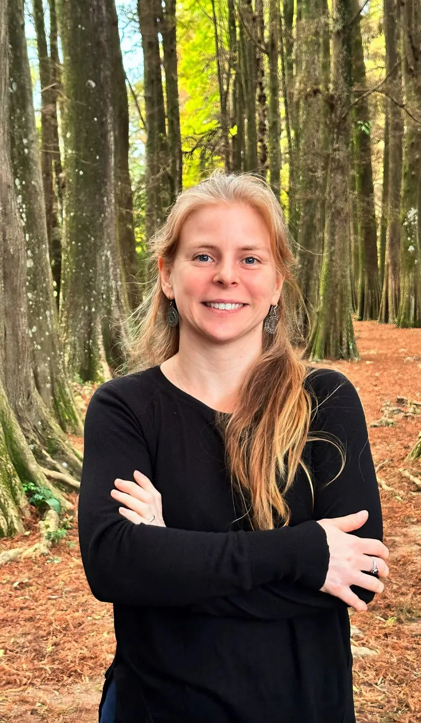 Retrato de Guta Adami em uma floresta, de braços cruzados e sorrindo
